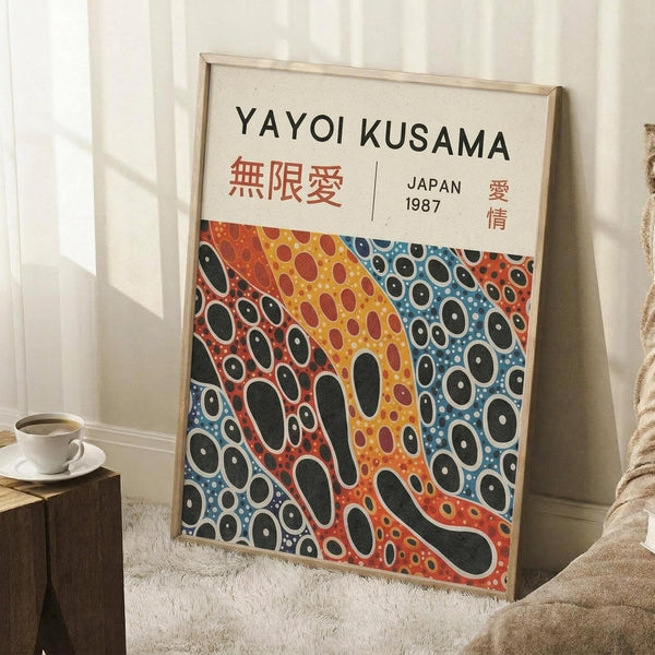 May include: Framed art print leaning against white wall corner on textured cream rug. Poster features Yayoi Kusama's name in black text with Japanese characters, dated Japan 1987. Artwork shows organic flowing shapes in vibrant orange, yellow, red and blue with black polka dots in Kusama's signature style. Light wood frame, cozy room setting with coffee cup on wooden block nearby and beige textiles.