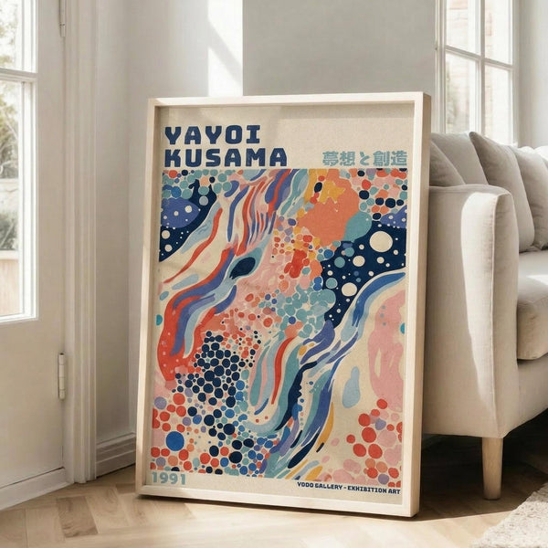 May include: Framed exhibition poster featuring abstract flowing design with coral, navy blue, and teal organic shapes, dots, and swirling patterns. Light wood frame leaning against white wall next to beige upholstered chair in bright, minimalist living room with large windows and natural lighting.