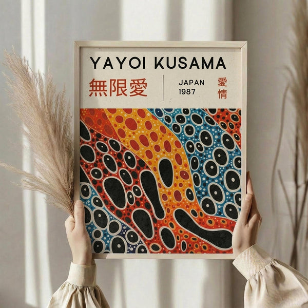 May include: Person holding framed poster with polka dot pattern in red, orange, yellow, and blue sections with black oval shapes throughout. Japanese text visible at top. Light wood frame against neutral interior with dried pampas grass and white curtains in soft natural lighting.