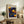 May include: Framed poster with blue background featuring yellow and orange polka dots in various sizes, resembling celestial bodies. Text reads "YAYOI KUSAMA" with Japanese characters and "KYOTO" below. Light wood frame sits on rustic wooden sideboard alongside fern plant, books, and ceramic vase in neutral-toned living room setting.
