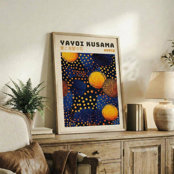 May include: Framed poster with blue background featuring yellow and orange polka dots in various sizes, resembling celestial bodies. Text reads "YAYOI KUSAMA" with Japanese characters and "KYOTO" below. Light wood frame sits on rustic wooden sideboard alongside fern plant, books, and ceramic vase in neutral-toned living room setting.