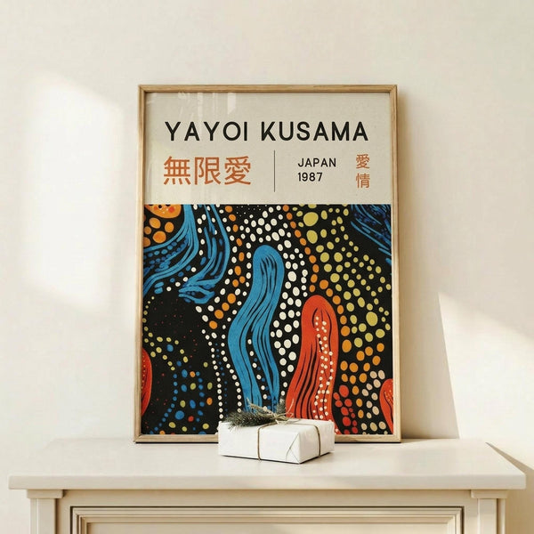 May include: Framed poster print featuring Yayoi Kusama-inspired artwork with vibrant polka dot patterns in blue, red, orange, and yellow against black background, flowing organic shapes, Japanese text, "JAPAN 1987" marking. Light wood frame displayed on minimal white wooden console table with small wrapped gift box beside it, white wall background.