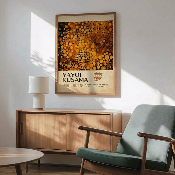 May include: A framed art print featuring vibrant orange and golden circular dots in Kusama's signature polka dot style, displayed above a wooden sideboard with a white ceramic lamp. The artwork shows "YAYOI KUSAMA" and Japanese characters on a cream background. A mint green mid-century armchair sits in the foreground with dramatic sunlight casting shadows across white walls.