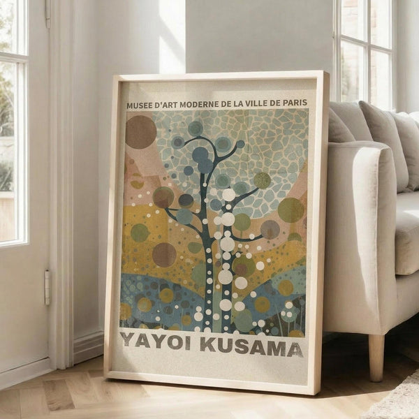 May include: Framed art print leaning against white wall next to beige upholstered chair in bright living room. Poster features abstract tree design with circular dots and organic shapes in muted green, yellow, blue and brown tones. Text reads "Musee d'Art Moderne de la Ville de Paris" at top and "Yayoi Kusama" at bottom. Light wooden frame, natural lighting from windows creates soft shadows on hardwood floors.