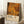 May include: A framed art print leaning against white walls on a textured cream rug. The poster features hundreds of golden, amber, and bronze circles in various sizes creating a dense pattern against a dark background. Below the circular design is cream-colored text reading "YAYOI KUSAMA" with Japanese characters and museum text. A white coffee cup sits on a wooden block nearby, with soft natural lighting creating shadows on the walls.