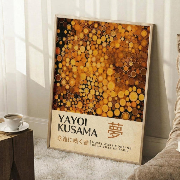 May include: A framed art print leaning against white walls on a textured cream rug. The poster features hundreds of golden, amber, and bronze circles in various sizes creating a dense pattern against a dark background. Below the circular design is cream-colored text reading "YAYOI KUSAMA" with Japanese characters and museum text. A white coffee cup sits on a wooden block nearby, with soft natural lighting creating shadows on the walls.