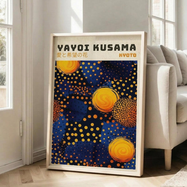 May include: A framed poster with Yayoi Kusama's signature polka dot style featuring three large yellow spiral orbs against a deep blue background filled with golden dots of varying sizes. The artwork is displayed in a light wooden frame leaning against a white wall next to a beige armchair with gray throw pillows. Natural sunlight streams through tall windows, creating a bright, modern living room setting on light wooden floors.