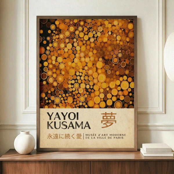 May include: Framed art poster displaying densely packed golden, orange, and amber circles of varying sizes against a dark background, with "YAYOI KUSAMA" text and Japanese characters below. The wooden-framed print sits on a mid-century modern credenza beside a white ceramic vase and books, against cream walls with white trim molding.