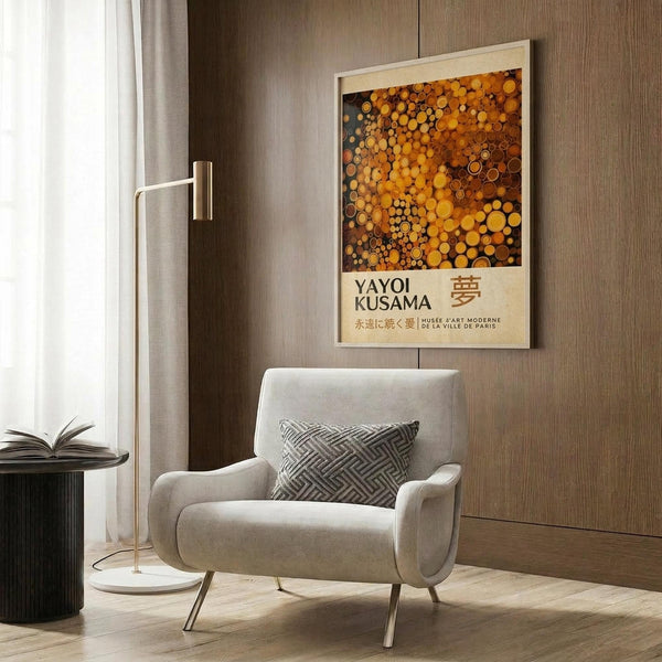 May include: Modern living room with beige armchair and geometric pillow, featuring framed art poster on wood-paneled wall. Artwork displays golden and orange circular dots pattern with Yayoi Kusama text and Japanese characters. Room includes brass floor lamp, dark side table with open book, white curtains, and warm wood flooring.