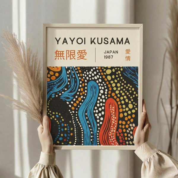May include: Person holding framed poster featuring abstract artwork with flowing blue and red organic shapes surrounded by colorful polka dots on black background. White matted frame with light wood border. Dried pampas grass stems visible on left side. Neutral beige curtains in soft indoor lighting create minimalist home decor setting.