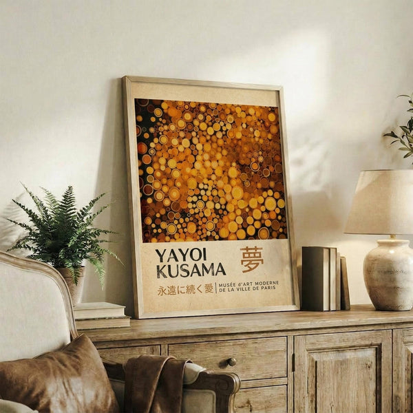 May include: Framed art print displaying colorful circular dots in warm orange, yellow and brown tones with Yayoi Kusama text below, positioned on rustic wooden sideboard alongside fern plant, ceramic vase, and books in neutral-toned living room with beige chair visible