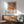 May include: Framed art poster featuring abstract tropical leaves in coral pink and yellow with black palm fronds and polka dot patterns, displayed above a wooden sideboard with white ceramic lamp in modern living room with mint green armchair and natural lighting casting shadows on white wall.
