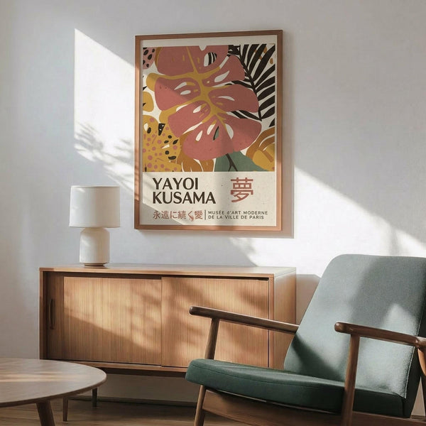 May include: Framed art poster featuring abstract tropical leaves in coral pink and yellow with black palm fronds and polka dot patterns, displayed above a wooden sideboard with white ceramic lamp in modern living room with mint green armchair and natural lighting casting shadows on white wall.