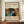 May include: Framed Yayoi Kusama exhibition poster displayed on wooden sideboard next to white ceramic vase and stacked books. The colorful artwork features abstract organic shapes with polka dots in vibrant blues, oranges, greens and reds against black background. Text shows "YAYOI KUSAMA" with Japanese characters and museum exhibition details. Cream matting surrounds the print in dark wooden frame against white paneled wall.