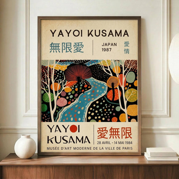 May include: Framed Yayoi Kusama exhibition poster displayed on wooden sideboard next to white ceramic vase and stacked books. The colorful artwork features abstract organic shapes with polka dots in vibrant blues, oranges, greens and reds against black background. Text shows "YAYOI KUSAMA" with Japanese characters and museum exhibition details. Cream matting surrounds the print in dark wooden frame against white paneled wall.