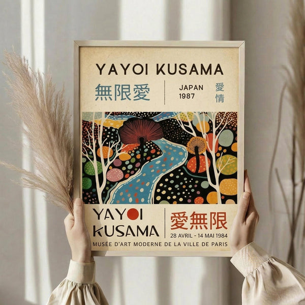 May include: Person holding framed exhibition poster in cream-colored frame against white curtained background, with dried pampas grass visible on left. Poster features colorful abstract artwork with polka-dotted organic shapes in blues, oranges, and earth tones, along with Japanese and English text including artist name and museum details.