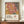 May include: Framed art poster with abstract botanical design featuring coral pink monstera leaf with yellow veins, black and white striped palm frond, and colorful spotted patterns. Text reads "YAYOI KUSAMA" with Japanese characters and "MUSÉE D'ART MODERNE DE LA VILLE DE PARIS." Displayed on wooden sideboard next to white ceramic vase and stacked books in bright room with white paneled walls.