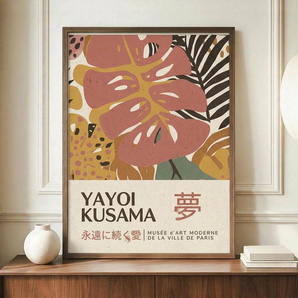 May include: Framed art poster with abstract botanical design featuring coral pink monstera leaf with yellow veins, black and white striped palm frond, and colorful spotted patterns. Text reads "YAYOI KUSAMA" with Japanese characters and "MUSÉE D'ART MODERNE DE LA VILLE DE PARIS." Displayed on wooden sideboard next to white ceramic vase and stacked books in bright room with white paneled walls.
