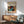 May include: Framed art poster featuring Yayoi Kusama's colorful abstract design with polka dots, organic shapes in blues, oranges, and earth tones, displayed above a wooden sideboard with white ceramic lamp. Mid-century modern teal upholstered chair with wooden arms sits beside round coffee table. Warm natural lighting creates shadows on white wall in minimalist living room setting.