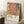 May include: Framed art poster featuring abstract botanical design with coral pink monstera leaf, yellow and black spotted elements, and striped palm fronds in warm earth tones. Text reads "YAYOI KUSAMA" with Japanese characters and "MUSÉE D'ART MODERNE DE LA VILLE DE PARIS". Poster leans against white wall corner next to wooden side table with white coffee cup and saucer, cream textured rug below, cozy natural lighting.