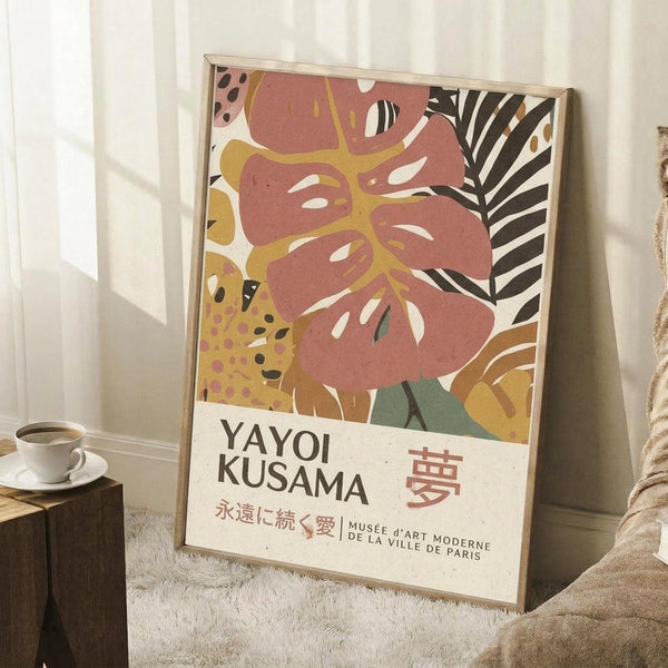 May include: Framed art poster featuring abstract botanical design with coral pink monstera leaf, yellow and black spotted elements, and striped palm fronds in warm earth tones. Text reads "YAYOI KUSAMA" with Japanese characters and "MUSÉE D'ART MODERNE DE LA VILLE DE PARIS". Poster leans against white wall corner next to wooden side table with white coffee cup and saucer, cream textured rug below, cozy natural lighting.