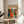 May include: Framed art print featuring vibrant abstract design with colorful polka-dotted organic shapes in orange, blue, red, and yellow on dark background, displayed on light wood round side table next to beige upholstered chair, with plant leaves visible in soft natural lighting against white wall