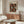 May include: Yayoi Kusama museum exhibition poster mockup displayed above mid-century modern walnut credenza in minimalist living room. Poster features repeating orange and white pumpkin motifs with black polka dot background in thin wooden frame. Scene includes cream sculptural chair, glass oval coffee table, and brass floor lamp against white walls.