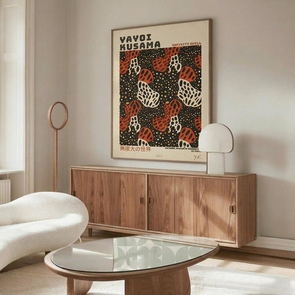 May include: Yayoi Kusama museum exhibition poster mockup displayed above mid-century modern walnut credenza in minimalist living room. Poster features repeating orange and white pumpkin motifs with black polka dot background in thin wooden frame. Scene includes cream sculptural chair, glass oval coffee table, and brass floor lamp against white walls.