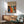 May include: A framed art print featuring abstract colorful patterns with dots, circles, and organic shapes in orange, blue, red, and yellow tones, displaying "YAYOI KUSAMA" and Japanese characters at the top with "JAPAN 1987". The wooden-framed poster hangs above a mid-century modern sideboard with a white ceramic table lamp, next to a mint green upholstered armchair with wooden frame. Warm sunlight creates geometric shadows on the white wall.