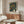 May include: Framed Yayoi Kusama exhibition poster with swirling green background and colorful dots displayed above a wooden sideboard in modern living room. Features warm wood credenza, white sculptural chair, glass coffee table, and copper floor lamp. Poster shows abstract organic shapes in teal, yellow, orange, and white circles with Japanese text and "TOKYO" at bottom.