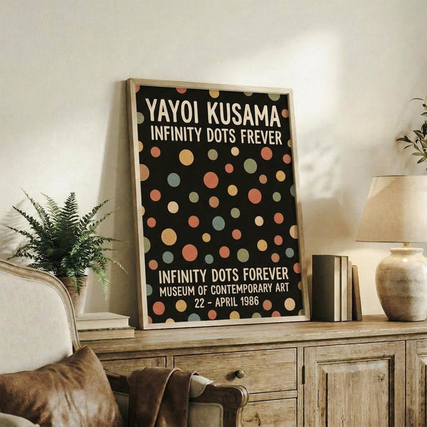 May include: Framed exhibition poster with black background and colorful polka dots in coral, sage green, cream, and peach tones, displayed on rustic wooden sideboard alongside fern plant, ceramic vase, and stacked books in neutral-toned living room setting