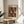 May include: Framed art print displayed on light wood round side table next to beige upholstered chair. Print features repeating pattern of red and white spotted mushrooms on dark background with small dots. Wooden frame with cream matting. Soft natural lighting creates shadows on white wall. Green plant leaves visible in corner of minimalist modern room setting.