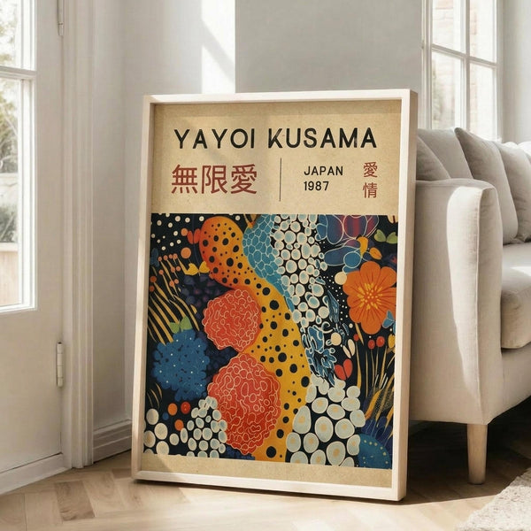 May include: Framed art print leaning against white wall next to beige upholstered chair in bright living room. Poster features colorful abstract pattern with polka dots, organic shapes, and floral motifs in orange, blue, red, and yellow on dark background. Top displays "YAYOI KUSAMA" text with Japanese characters and "JAPAN 1987". Natural wood frame with cream matting, approximately 2-3 feet tall.