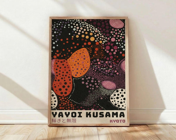 May include: Framed poster with Yayoi Kusama-inspired polka dot artwork featuring vibrant orange, pink, purple and white circles of varying sizes against a black background, displayed in light wood frame leaning against white wall on light wooden floor with soft natural lighting casting shadows