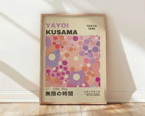 May include: Framed poster with cream background featuring colorful overlapping circles in purple, pink, orange, and coral tones. Text reads "YAYOI KUSAMA" in purple and black letters, "TOKYO 1998" in upper right, and Japanese characters at bottom with "22 APRIL 1986" and "MUSEE D'ART MODERNE DE LA VILLE DE PARIS." Natural wood frame sits on light wooden floor against white wall with soft shadows.
