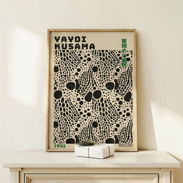 May include: A framed art poster with black polka dot pattern on cream background, featuring "YAYOI KUSAMA" text at top and "1992" at bottom with green accent text. The poster is displayed in a light wood frame on a pale wooden shelf against a white wall, with a small wrapped white gift box beside it.