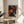 May include: Framed colorful abstract art print featuring vibrant polka dot patterns in orange, pink, white, and black with organic flowing shapes, displayed on a light wood round side table next to a cream upholstered chair. Soft natural lighting creates gentle shadows on white walls, with a plant visible in the corner.