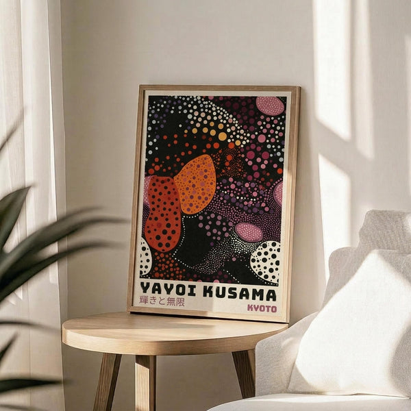 May include: Framed colorful abstract art print featuring vibrant polka dot patterns in orange, pink, white, and black with organic flowing shapes, displayed on a light wood round side table next to a cream upholstered chair. Soft natural lighting creates gentle shadows on white walls, with a plant visible in the corner.