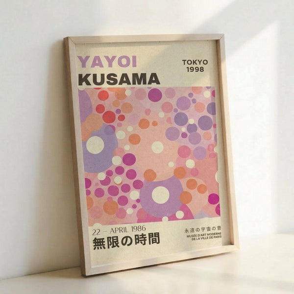 May include: Framed exhibition poster featuring colorful polka dot pattern in pink, purple, orange and cream circles of varying sizes. Light wood frame leaning against white wall. Japanese text at bottom reads "22 - APRIL 1986" and "無限の時間". Artist name "YAYOI KUSAMA" and "TOKYO 1998" printed at top, with "MUSÉE D'ART MODERNE DE LA VILLE DE PARIS" referenced below.