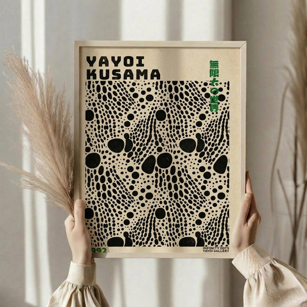 May include: Person in cream sweater holding framed art print featuring black polka dot pattern on beige background, with "YAYOI KUSAMA" text at top and green Japanese characters. Light wood frame, pampas grass visible to left, soft natural lighting against neutral curtained background.