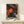 May include: Framed poster with colorful dotted abstract design in orange, pink, white and black polka dot patterns on dark background, displayed on light wood surface next to small wrapped gift box. Artwork features flowing organic shapes filled with various sized circles creating psychedelic visual effect. Natural lighting casts soft shadows on cream wall behind minimal wooden frame.