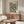 May include: Framed Yayoi Kusama exhibition poster with black polka dot pattern on cream background, displayed above mid-century walnut credenza in minimalist living room. White sculptural chair and oval glass coffee table complete the scene. Poster features bold typography and 1992 date marking.