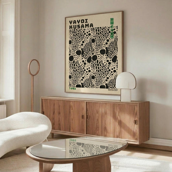 May include: Framed Yayoi Kusama exhibition poster with black polka dot pattern on cream background, displayed above mid-century walnut credenza in minimalist living room. White sculptural chair and oval glass coffee table complete the scene. Poster features bold typography and 1992 date marking.