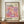 May include: Framed art print with colorful polka dot pattern in pink, purple, orange and cream tones, featuring "YAYOI KUSAMA" and Japanese text, displayed on wooden sideboard next to white ceramic vase and stacked books in cream-colored room with white paneled walls.