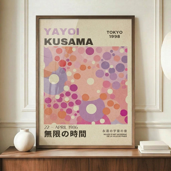 May include: Framed art print with colorful polka dot pattern in pink, purple, orange and cream tones, featuring "YAYOI KUSAMA" and Japanese text, displayed on wooden sideboard next to white ceramic vase and stacked books in cream-colored room with white paneled walls.