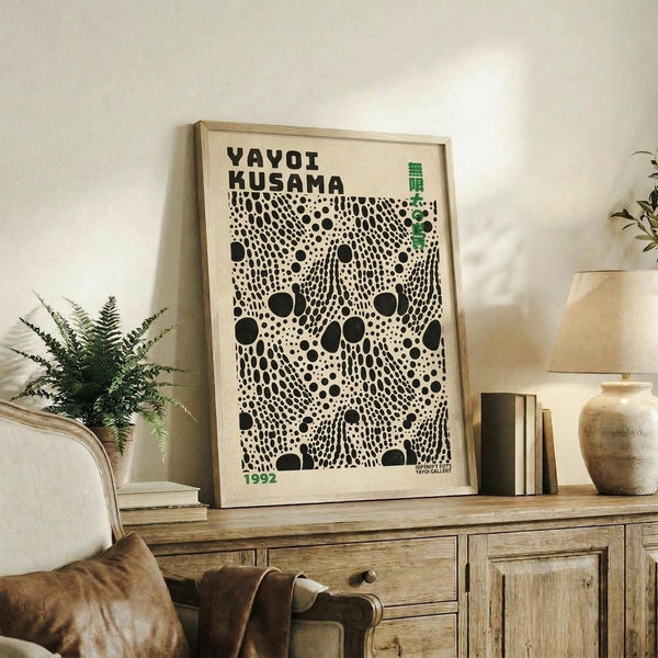 May include: Framed poster with black polka dot pattern on cream background, displayed on rustic wooden sideboard alongside fern plant, ceramic vase, and stacked books in neutral-toned living room setting with beige upholstered chair visible in foreground.