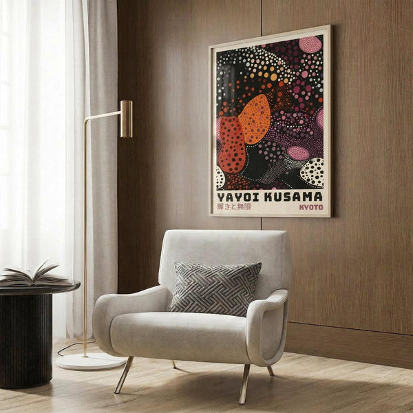 May include: Colorful abstract art poster with polka dot patterns in orange, pink, white, and black circular shapes, displayed in light wood frame on wood panel wall. Modern beige armchair with geometric patterned pillow sits below artwork. Gold floor lamp and dark round side table with open book complete the contemporary room setting with neutral tones and natural lighting.