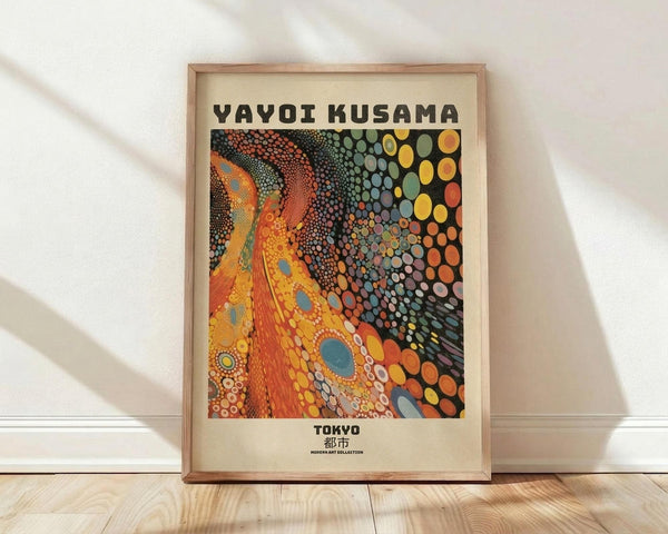 May include: Framed poster featuring Yayoi Kusama's distinctive polka dot artwork in vibrant oranges, yellows, and multicolored circular patterns against cream matting. Natural wood frame sits on light wooden floor against white wall with soft shadows from window light. The artwork shows flowing organic forms covered in signature infinity dots transitioning from warm tones to cooler blues and greens.