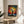 May include: Framed art print featuring a vibrant tree with bare black branches against a cosmic background of colorful dots in rainbow hues from red to blue. The artwork sits on a light wood round side table next to a beige upholstered chair, with a potted plant visible in soft natural lighting. The piece has Japanese text below the tree illustration and appears to be styled in a modern living room setting.