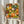 May include: Person holding framed art print featuring colorful dotted floral pattern in orange, green, yellow, and white circles against dark background. Japanese text visible at bottom. Natural lighting with dried wheat stalks and neutral curtains in background. Hands wearing cream colored ruffled sleeves grip wooden frame edges.